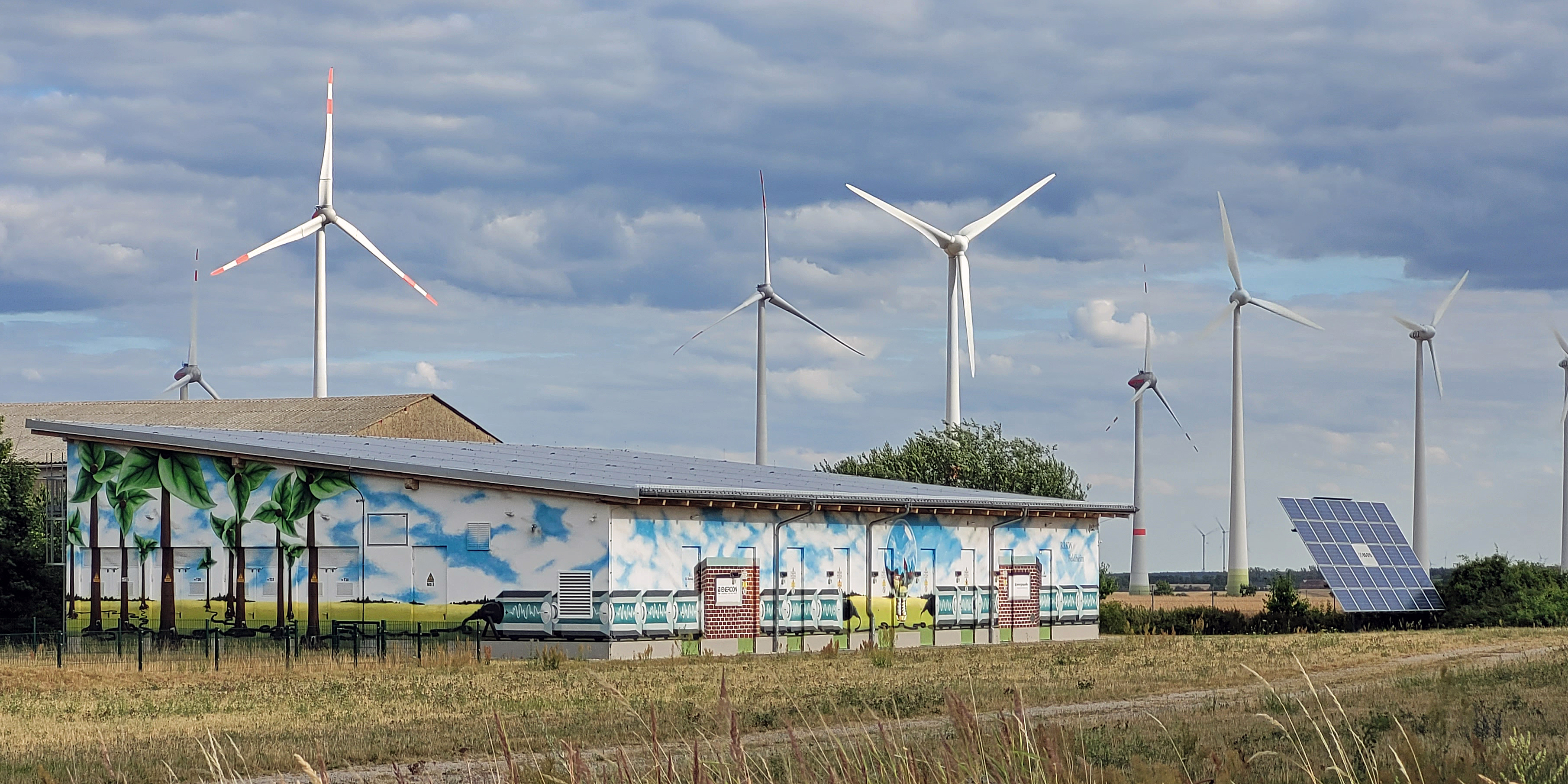 Windkraftanlanlagen, eine Solaranlage und ein flaches Gebäude, in dem elektrische Energie gespeichert wird. Die Fassade ist farbig gestaltet.
