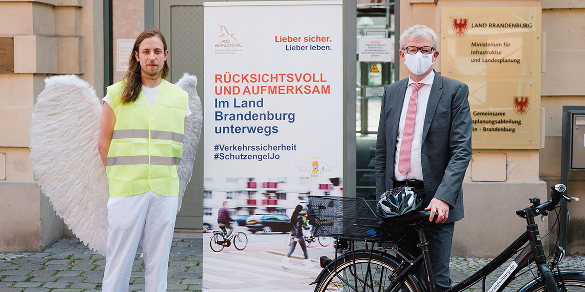 Minister Beermann und Kampagnen-Schutzengel Jo Ein Mann mit Schutzengel-Kostüm und ein Mann mit Fahrrad stehen vor einer Werbewand