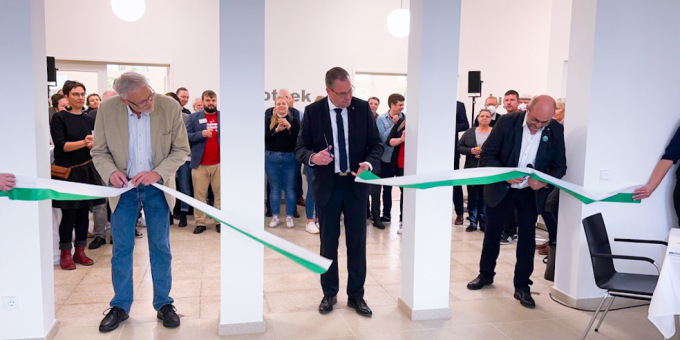 Staatssekretär Rainer Genilke durchschneidet ein grün-weißes Band im Hintergrund Teilnehmende der Eröffnungsveranstaltung