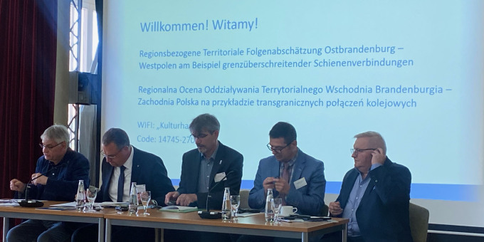 Podium der Auftaktveranstaltung, im Hintergrund die Präsentation
