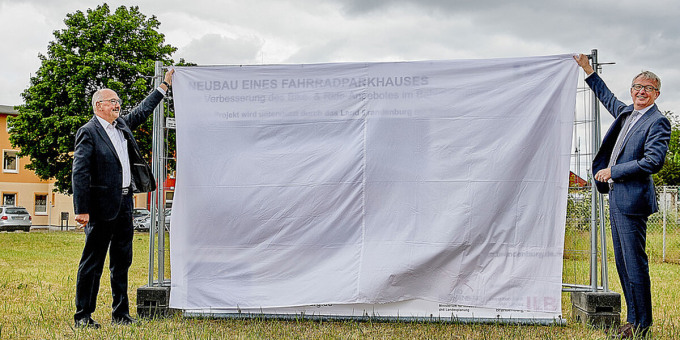 Minister Guido Beermann neben dem verhüllten Schild mit den Bauprojektinformationen